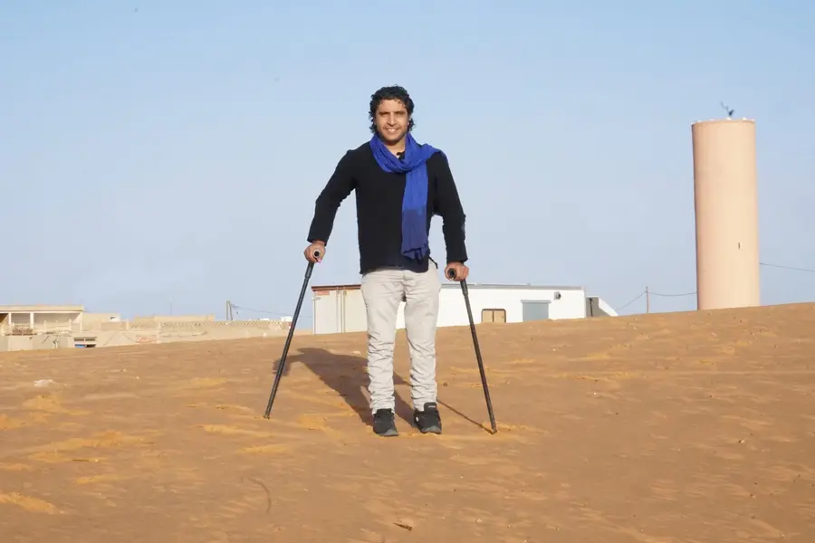 A smiling man with curly dark hair stands using two forearm crutches on a sandy dune, wearing a black top with a blue scarf and light gray pants. Behind him is a low town with flat-roofed buildings and a tall cylindrical water tower, under a clear pale blue sky