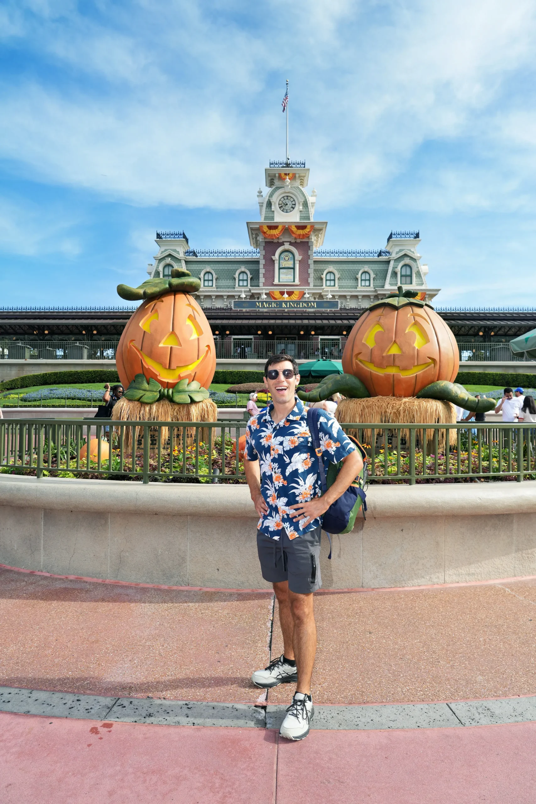Man standing outside the Magic Kingdom train station between two giant decorative pumpkin scarecrows.