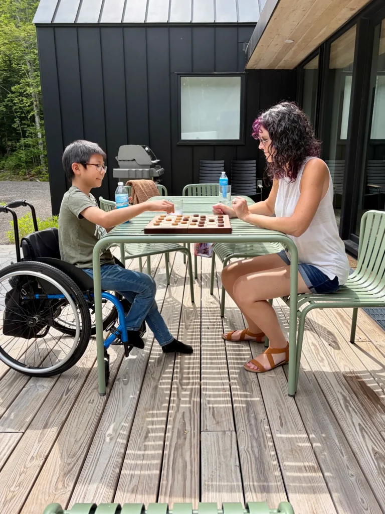 A young boy sitting in a wheelchair plays a board game at a green outdoor table on a wooden deck with a woman.