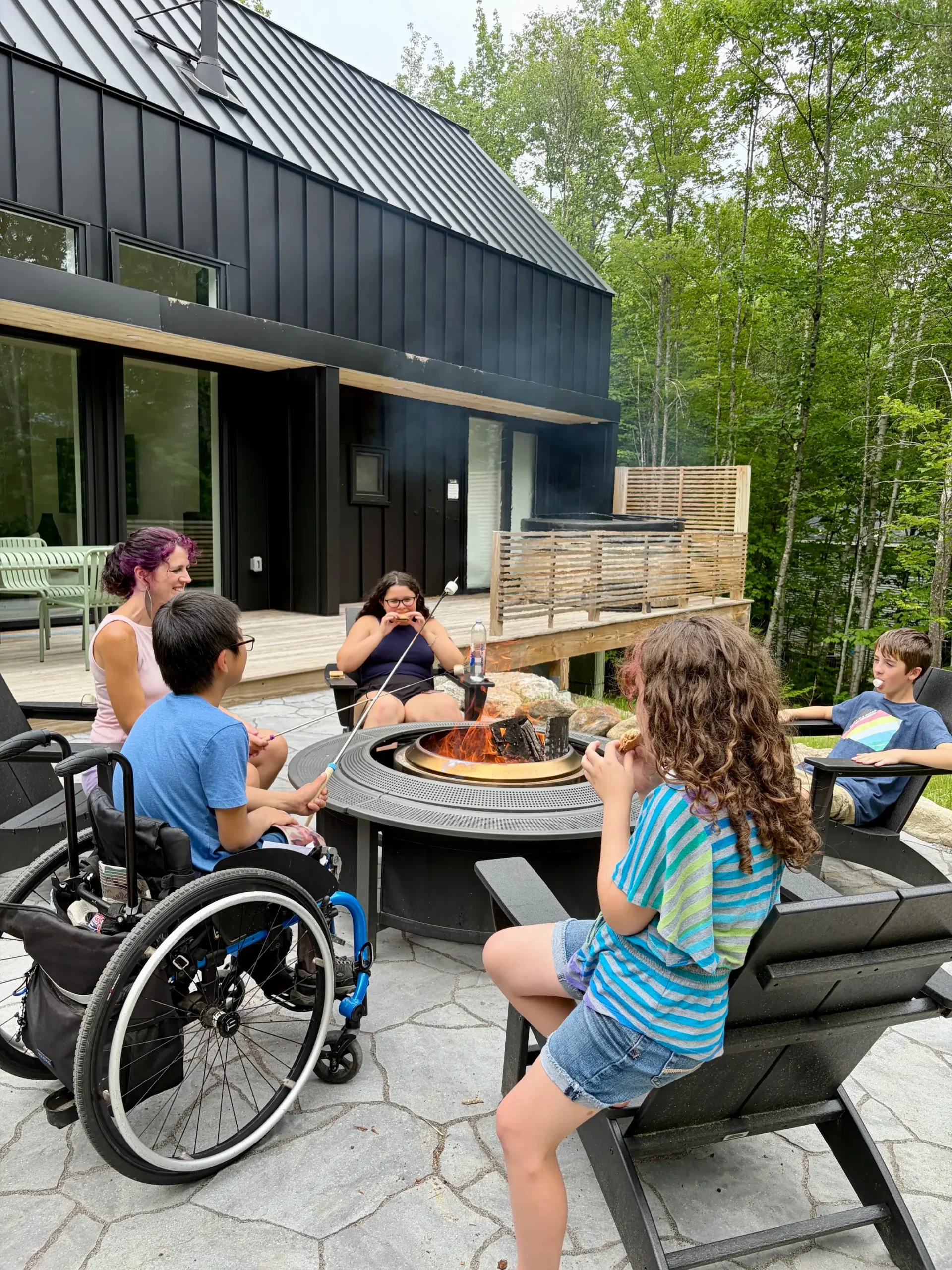 A group of adults and children gather around a modern outdoor fire pit roasting marshmallows outside a black modern house surrounded by trees.
