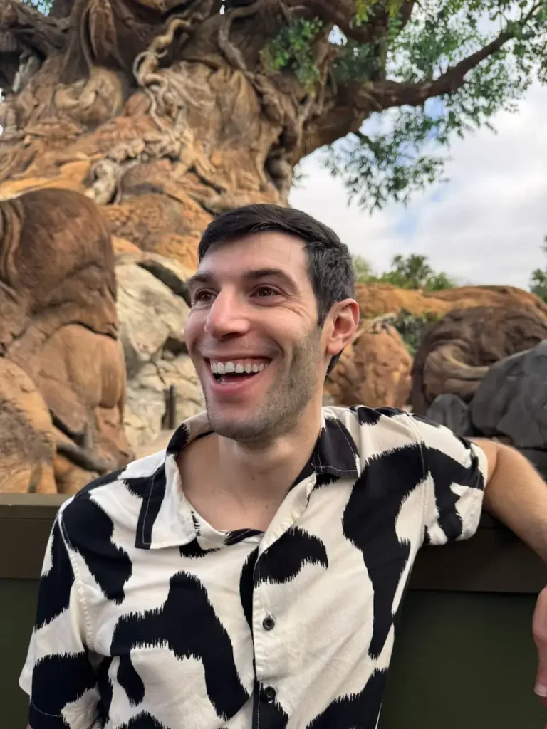 Smiling man in a black-and-white patterned button-down shirt standing outdoors in front of a large carved wooden tree.