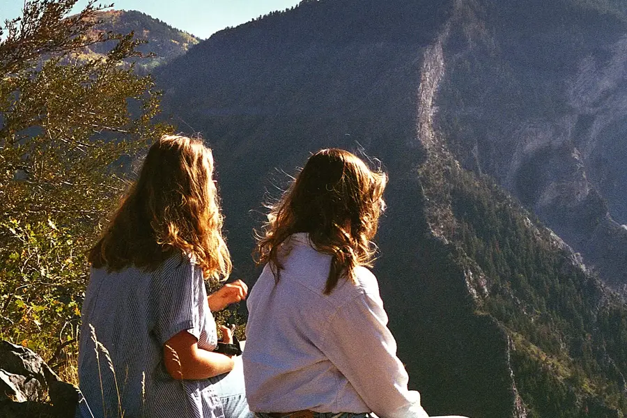 Two people seen from behind, sitting on a mountainside and gazing out at a dramatic, sunlit mountain landscape.