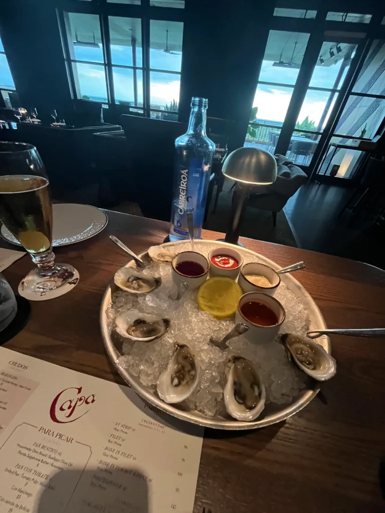 Platter of fresh raw oysters served on crushed ice with dipping sauces, lemon, and a bottle of Cabreiroá mineral water on a restaurant table.