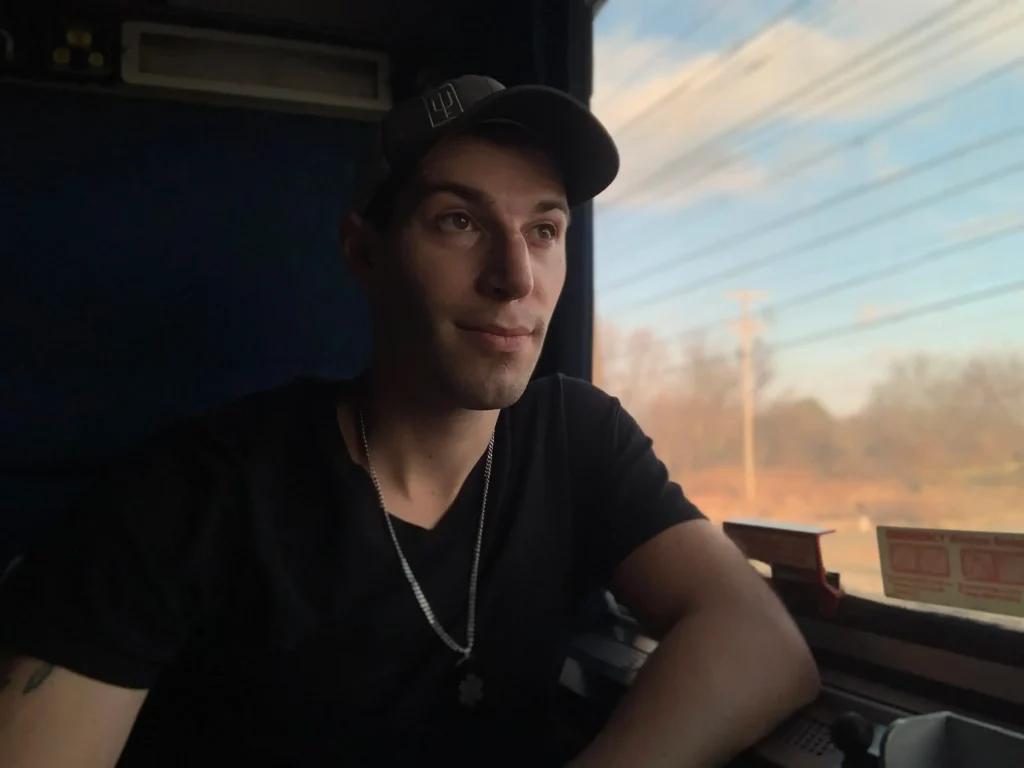 Man wearing a black baseball cap and t-shirt sitting by a window aboard a bus or train during golden hour.