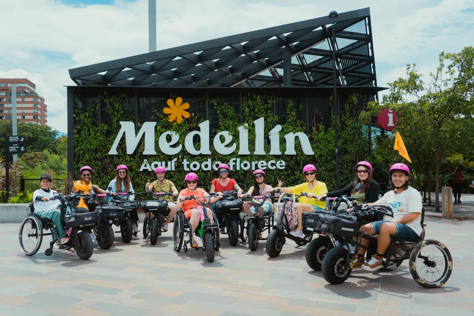 A group of people wearing pink helmets and riding electric handcycle wheelchairs lined up in front of a large "Medellín" sign.