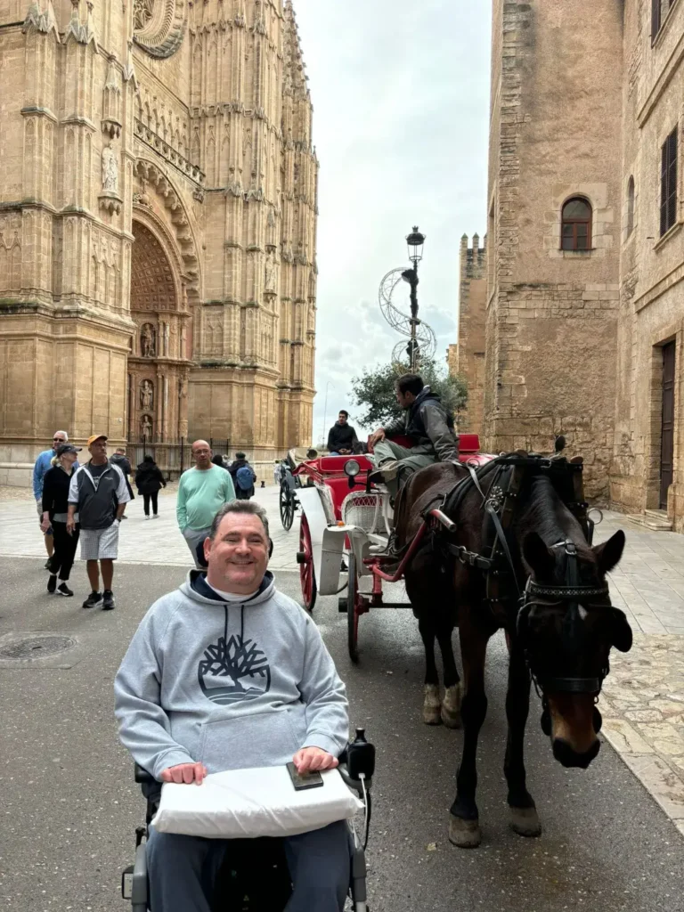 A man smiling in a wheelchair on a paved street beside a horse-drawn carriage, with the towering gothic facade of Palma Cathedral in the background.