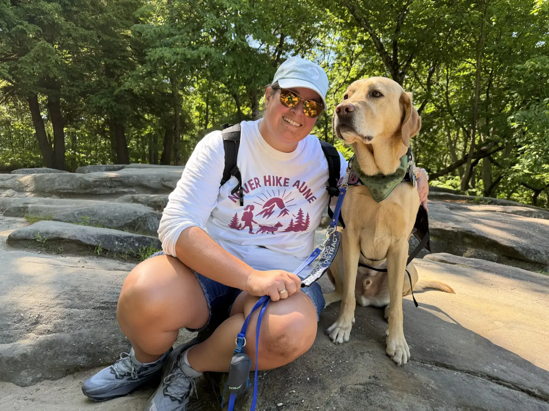 A smiling person wearing a cap, sunglasses, and a "Never Hike Alone" shirt sitting on large rocks next to their yellow Labrador guide dog in a sunny forest.