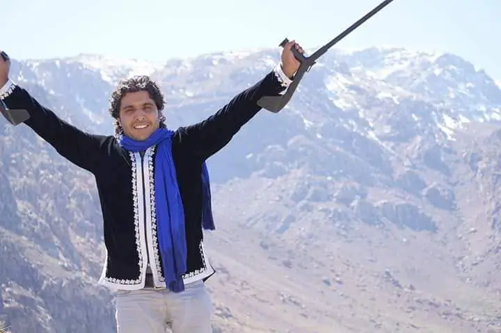 A smiling man holding his crutches high in the air in celebration, wearing a blue scarf against a rugged mountain landscape.