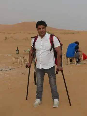 A smiling man wearing a backpack stands on desert sand holding forearm crutches, with dunes visible behind him.