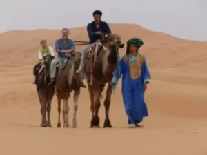 A camel caravan carrying tourists across sand dunes, led by a local guide in traditional blue robes.