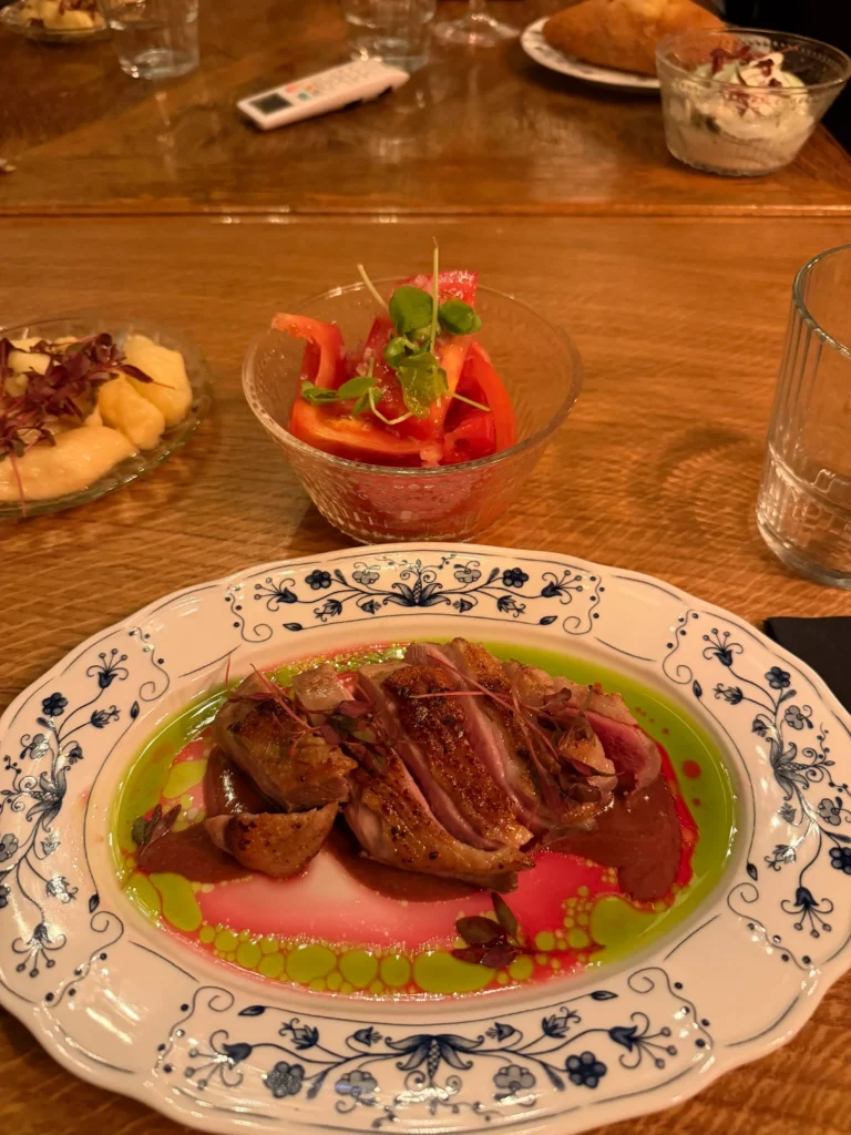 Sliced seared duck breast served over rich demi-glace and herb oil, accompanied by a small bowl of sliced fresh tomatoes.