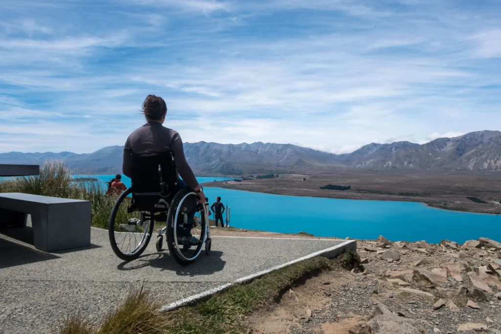 Rear view of a person using a manual wheelchair on a concrete lookout, gazing across a bright turquoise lake and distant mountain range.