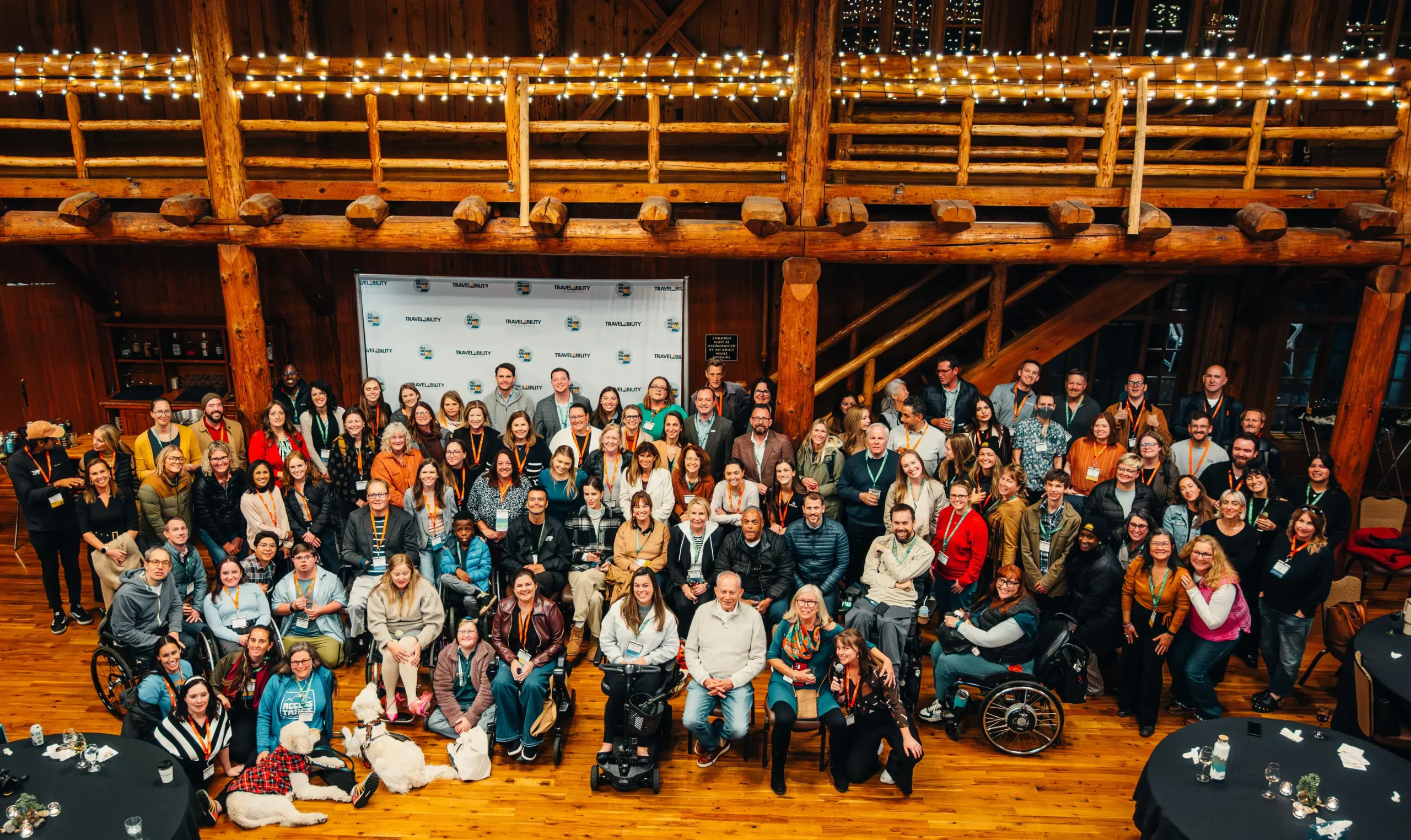A large group of attendees posing together inside a rustic wooden lodge decorated with string lights.