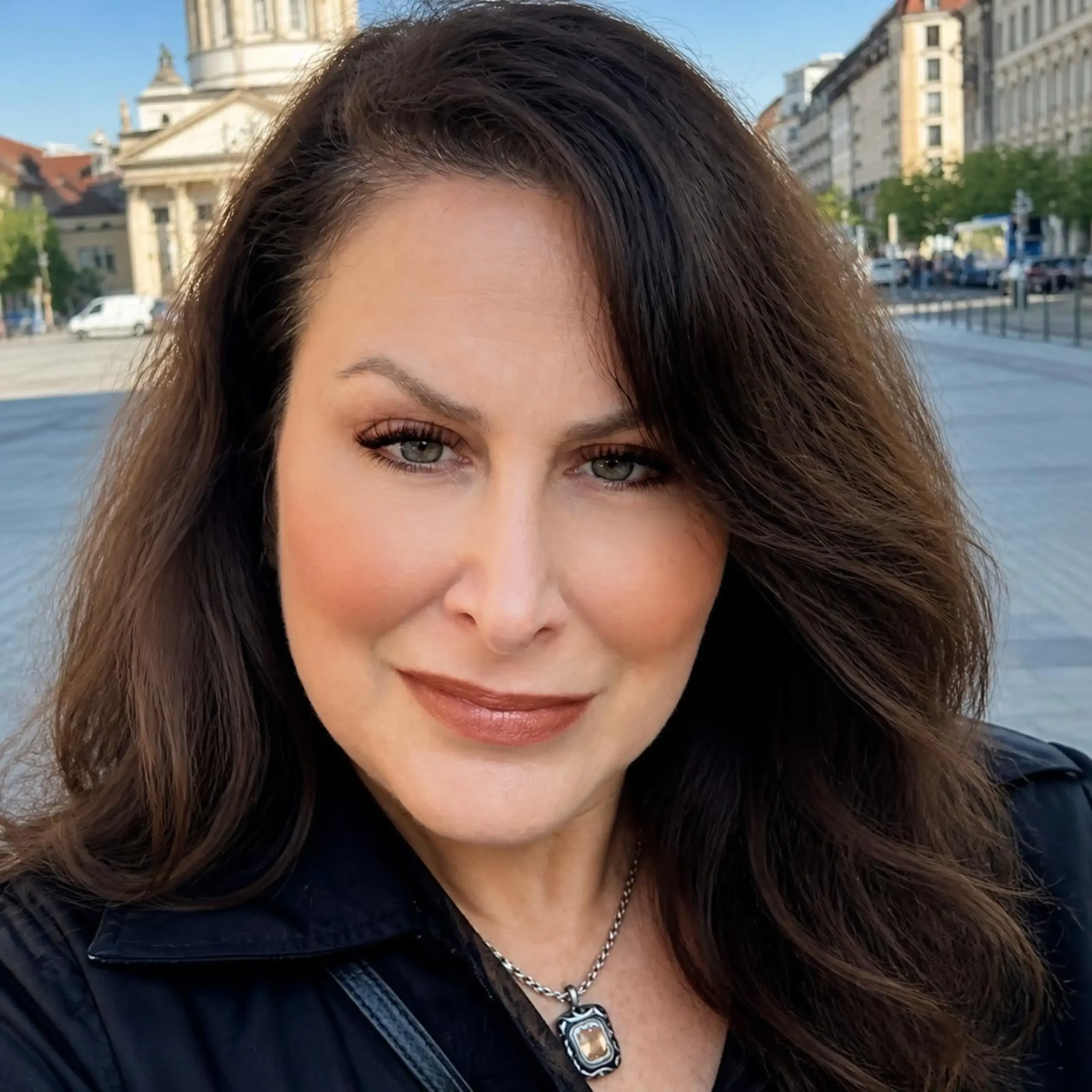 Close-up outdoor portrait of a smiling woman with long wavy brunette hair in a city square.