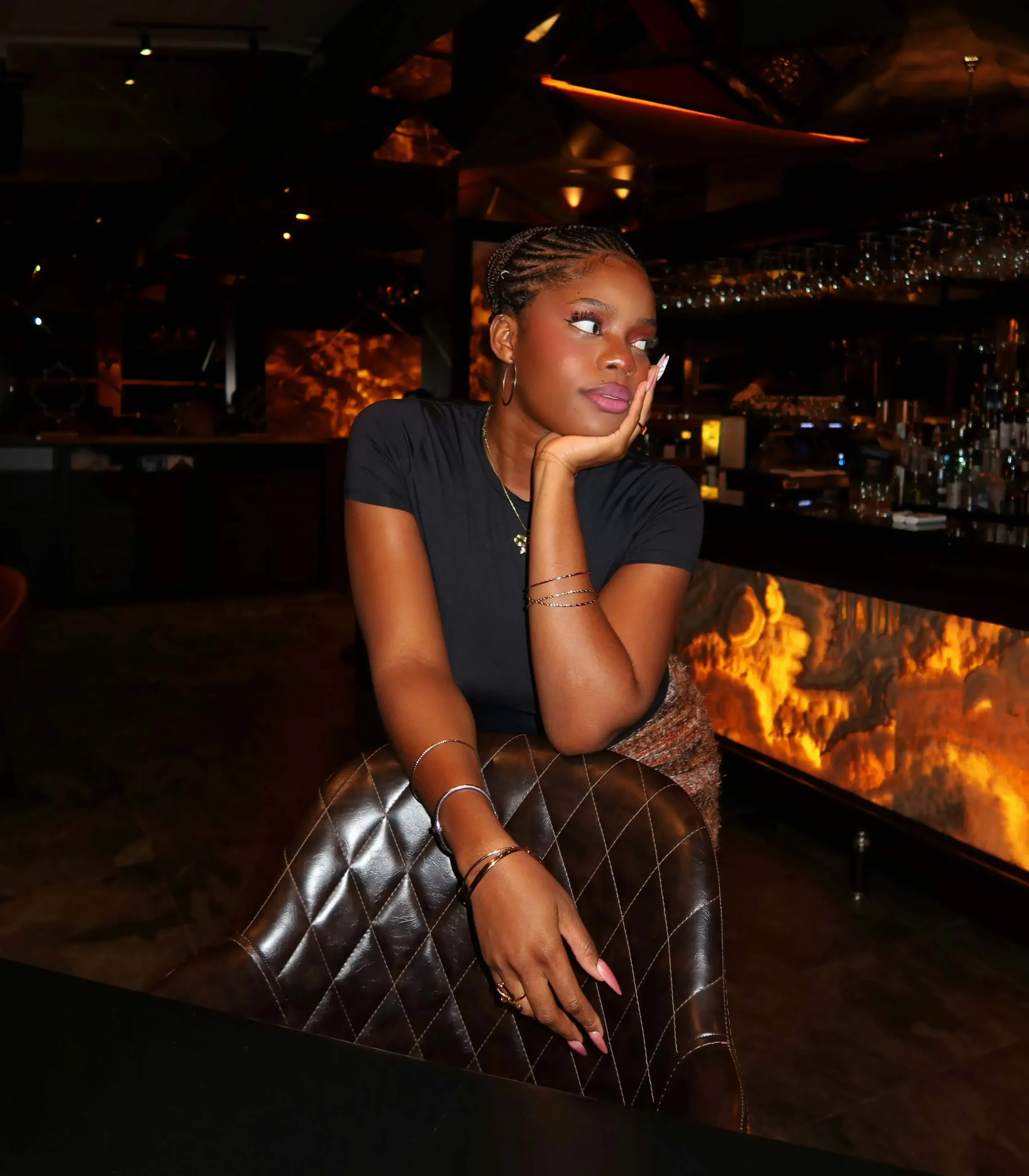Young woman leaning on a leather chair in a dimly lit bar with an illuminated counter.