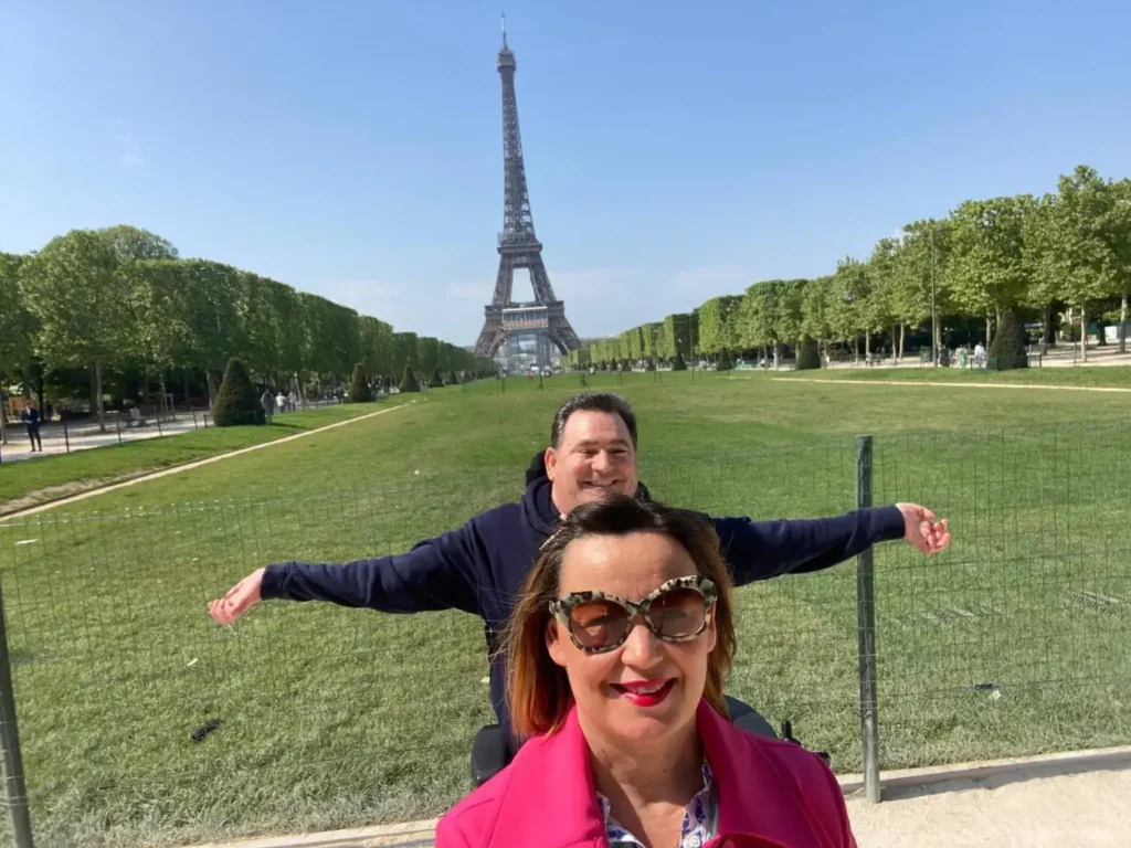 A smiling man in a wheelchair with arms outstretched behind a smiling woman wearing sunglasses and a pink coat, with a green lawn and the Eiffel Tower behind them.