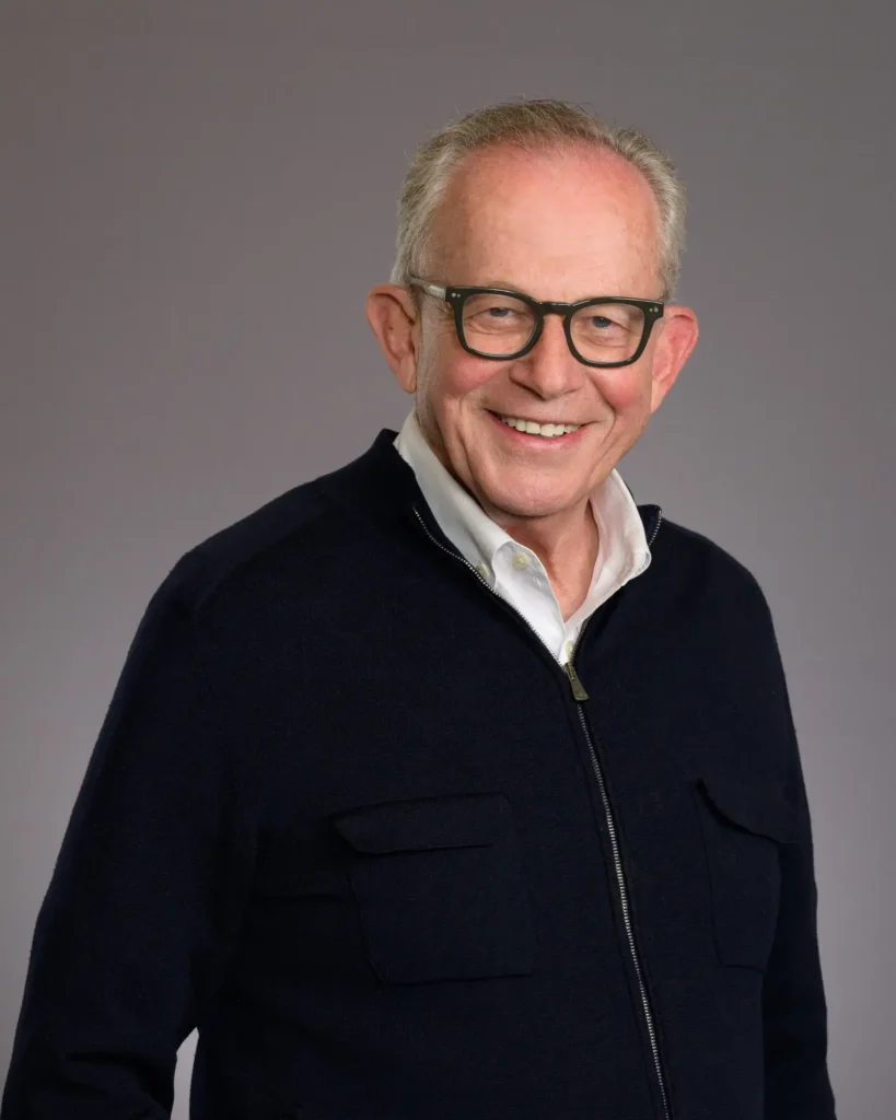 Portrait of an older man with gray hair and glasses smiling warmly, wearing an open-collar white shirt under a navy blue cardigan.