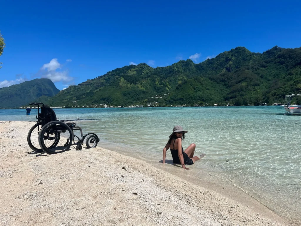 A woman in a swimsuit and sun hat sits in the shallow water of a sandy beach next to an all-terrain wheelchair, with lush green tropical mountains in the background.