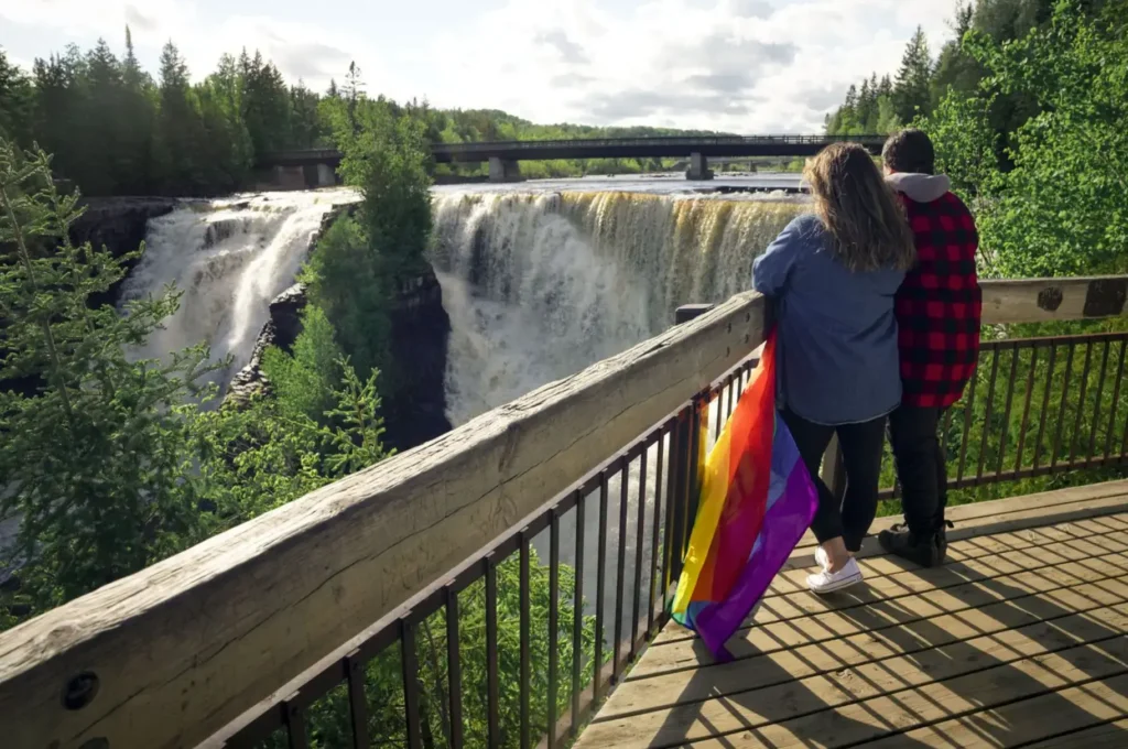 Two people standing at a wooden observation deck overlooking a large waterfall, with a pride flag draped over the railing beside them.