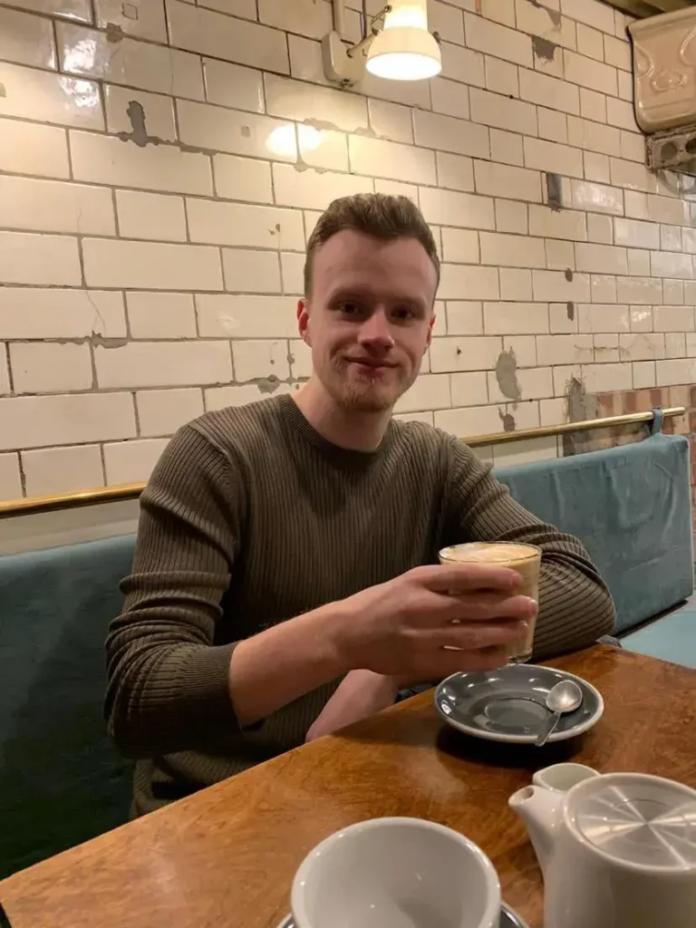 Louie Amos smiling while holding a glass of iced coffee at a wooden cafe table.