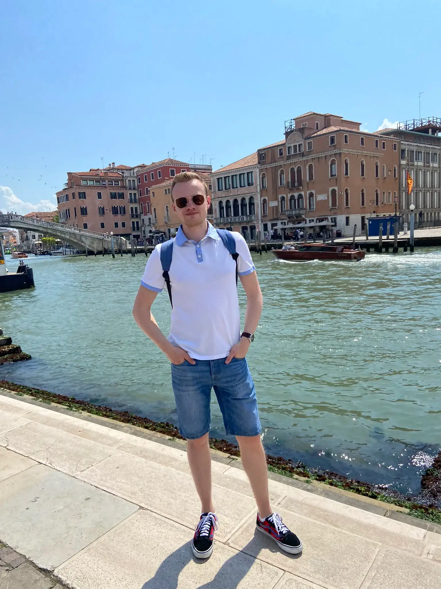 Louie Amos wearing sunglasses, a white polo shirt, and denim shorts while standing by a canal in Venice.