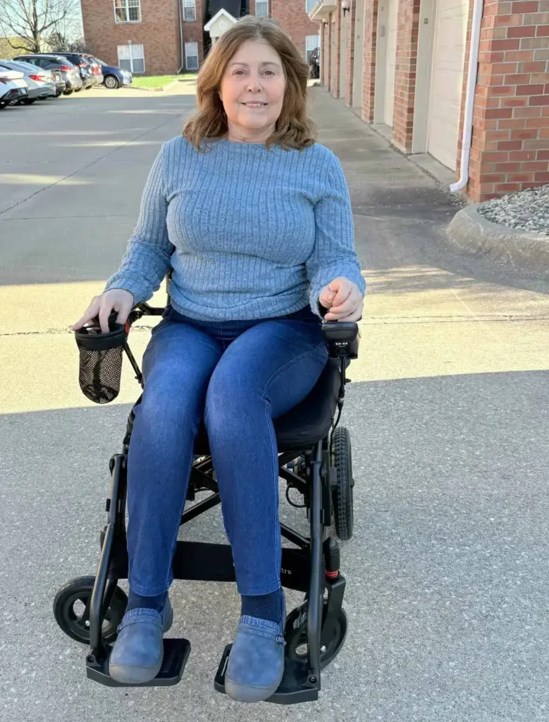 Marcia Frost smiling while seated in an electric power wheelchair outdoors on a paved path.