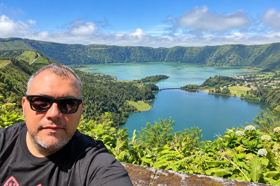 A man wearing sunglasses and a black t-shirt takes a selfie from a scenic overlook, with a panoramic view behind him of twin crater lakes—one turquoise, one deep blue—nestled between lush green hills, with a small village visible along the shoreline.
