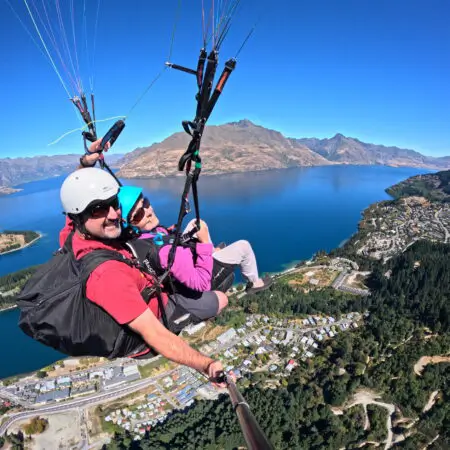 Two people tandem paragliding high above a deep blue lake, surrounding mountains, and a lakeside town.
