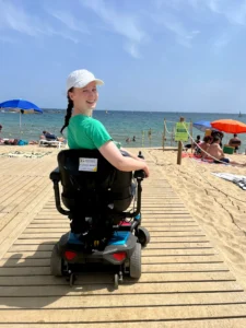 Woman in a green shirt and white cap smiling from a motorized wheelchair on a wooden beach walkway leading to the sea.