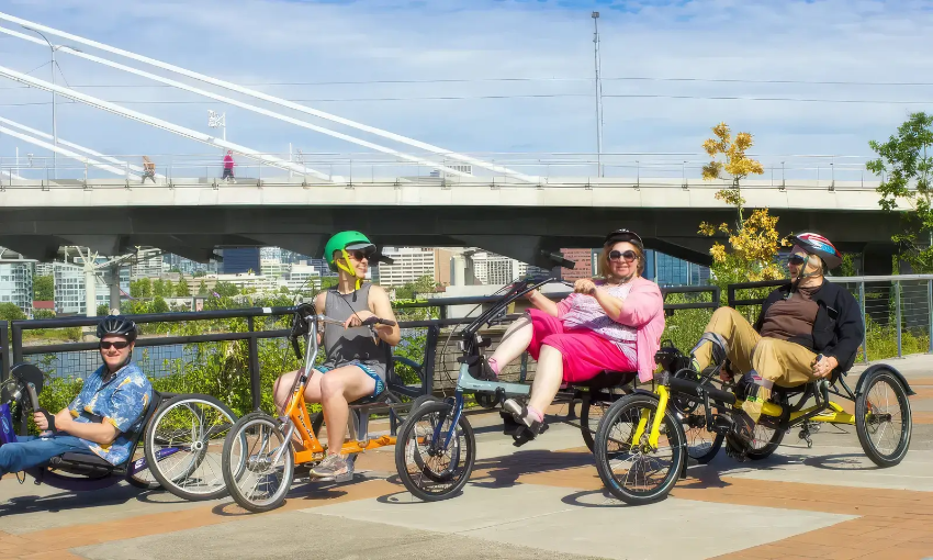 Four smiling cyclists riding recumbent bicycles and adaptive trikes along a paved waterfront path.