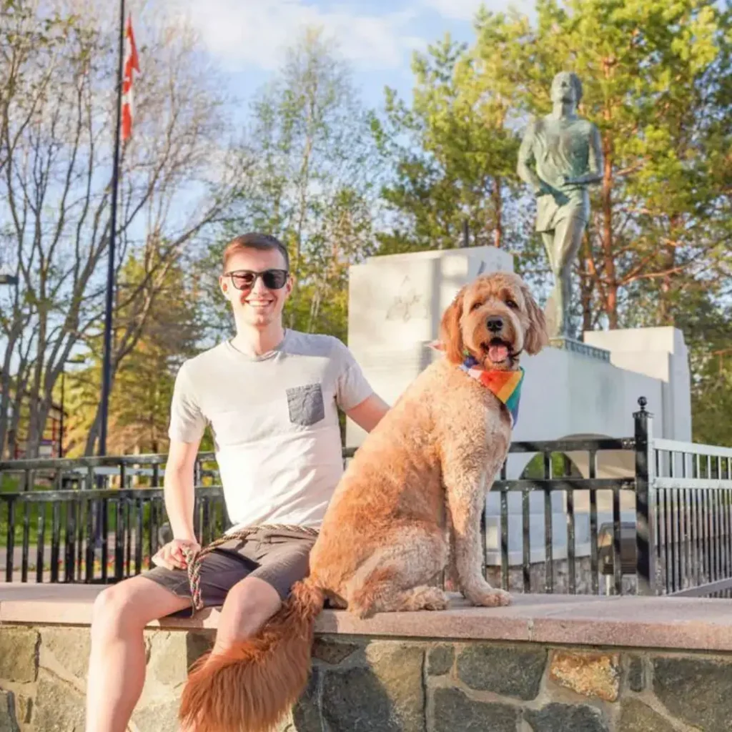 A smiling man in sunglasses sitting on a stone ledge beside a large golden dog wearing a rainbow bandana, with a running statue in the background.