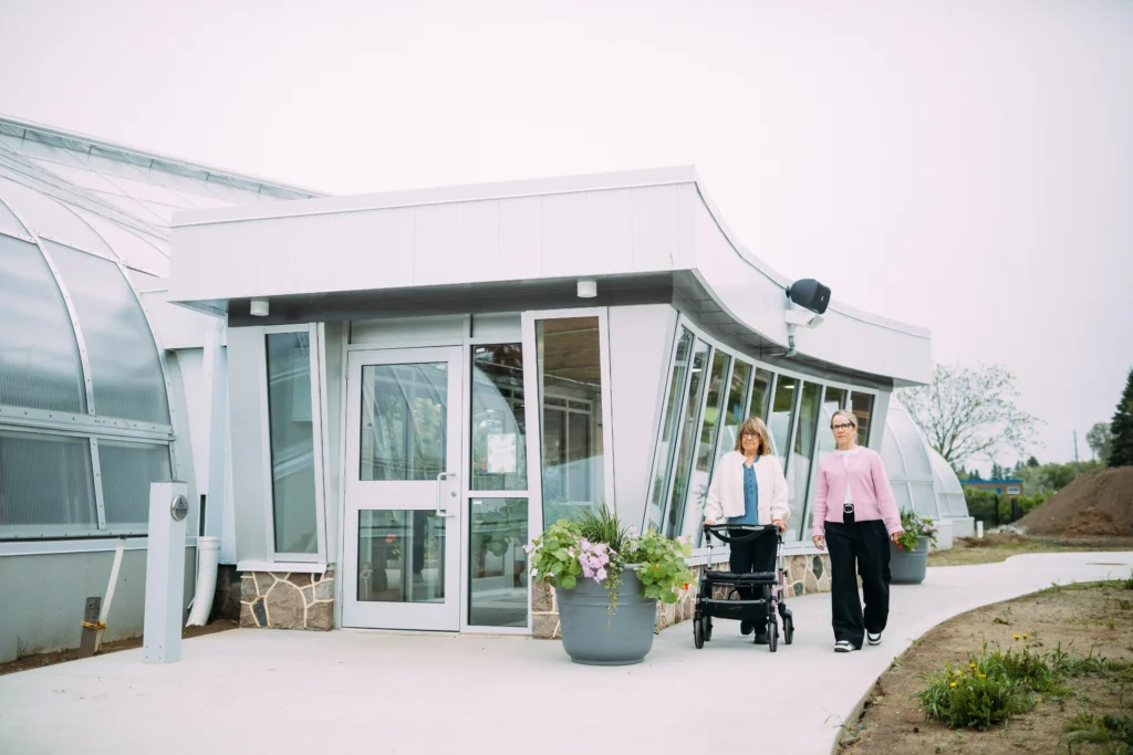 Two women walking side-by-side along a paved path outside a modern glass greenhouse, one using a wheeled walker.