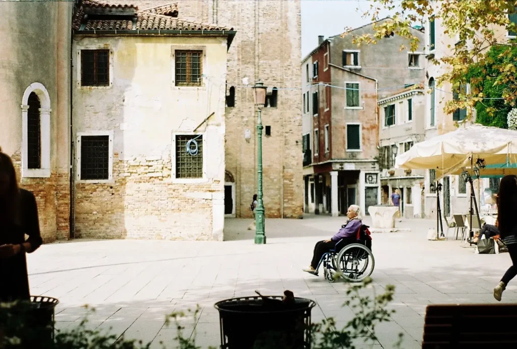 An older person in a wheelchair sits in a sunny cobblestone square surrounded by historic Italian buildings.