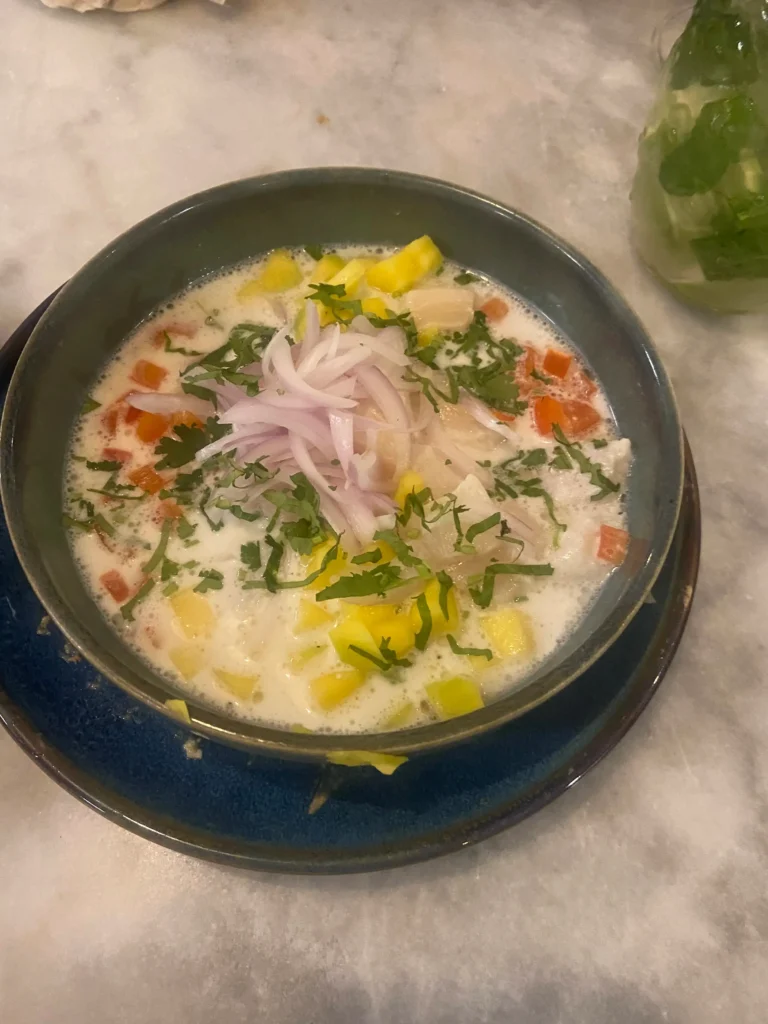 Bowl of white fish ceviche in creamy leche de tigre topped with red onion slices, diced mango, and fresh cilantro.
