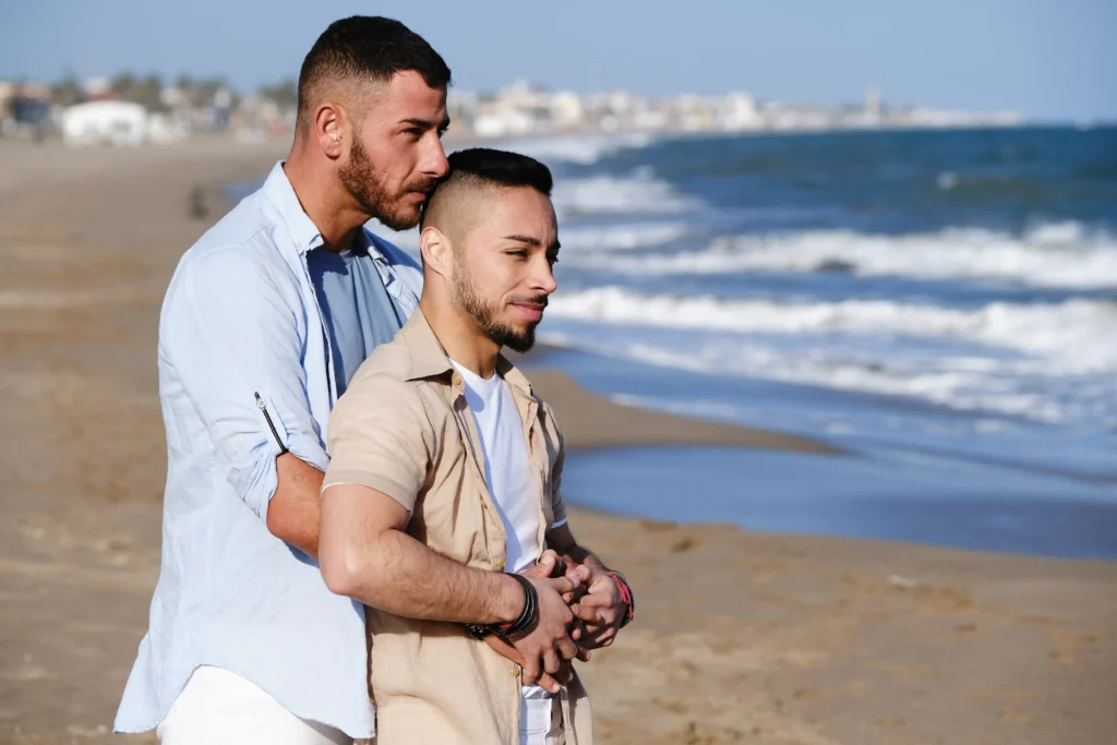 Two men embracing warmly while standing together on a sandy beach near the ocean shore.