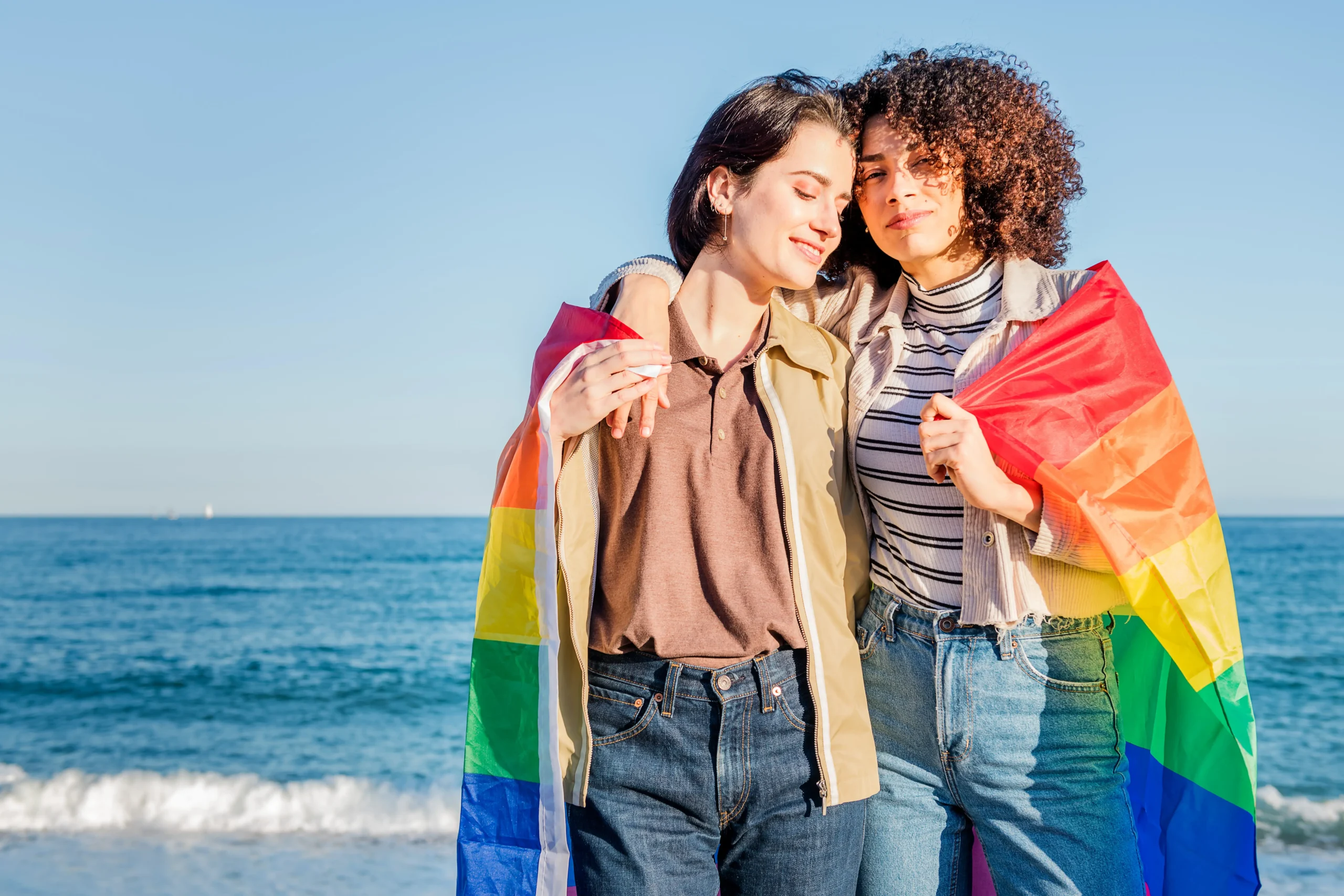 Two women smiling and embracing on a sunny beach draped in a vibrant rainbow pride flag.