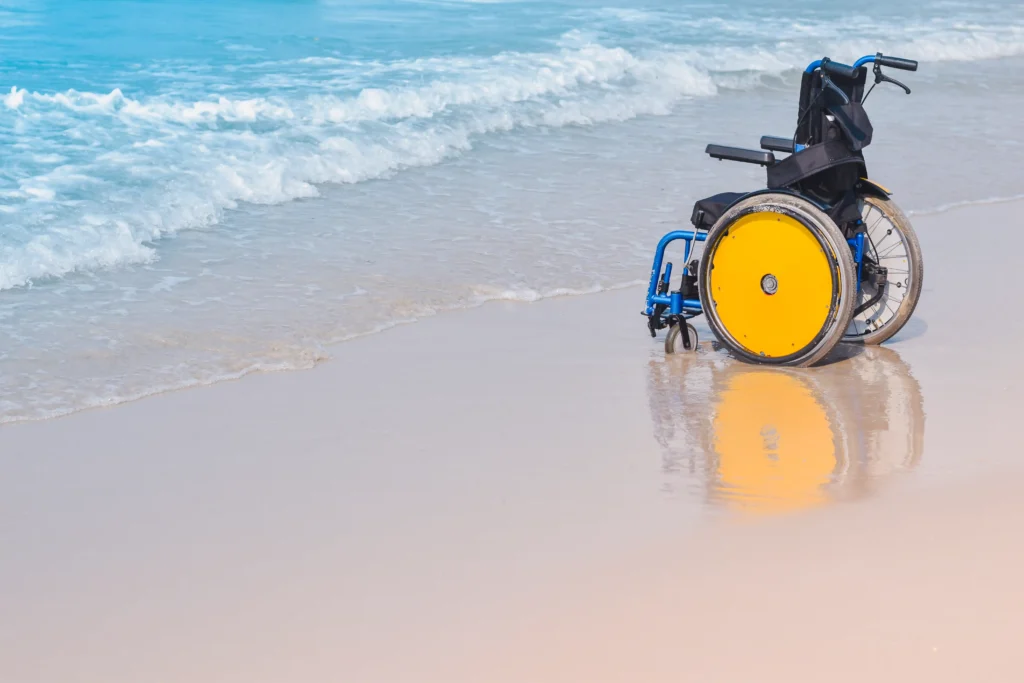 An empty blue and yellow wheelchair resting on wet sand at the edge of gentle ocean waves.